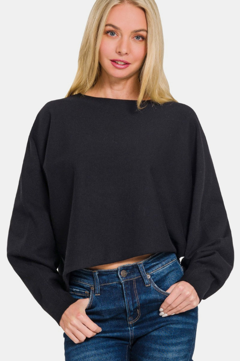 A woman with long blonde hair is wearing a Zenana Asymmetric Hem Long Sleeve Sweater in black paired with blue jeans, standing against a plain white background.