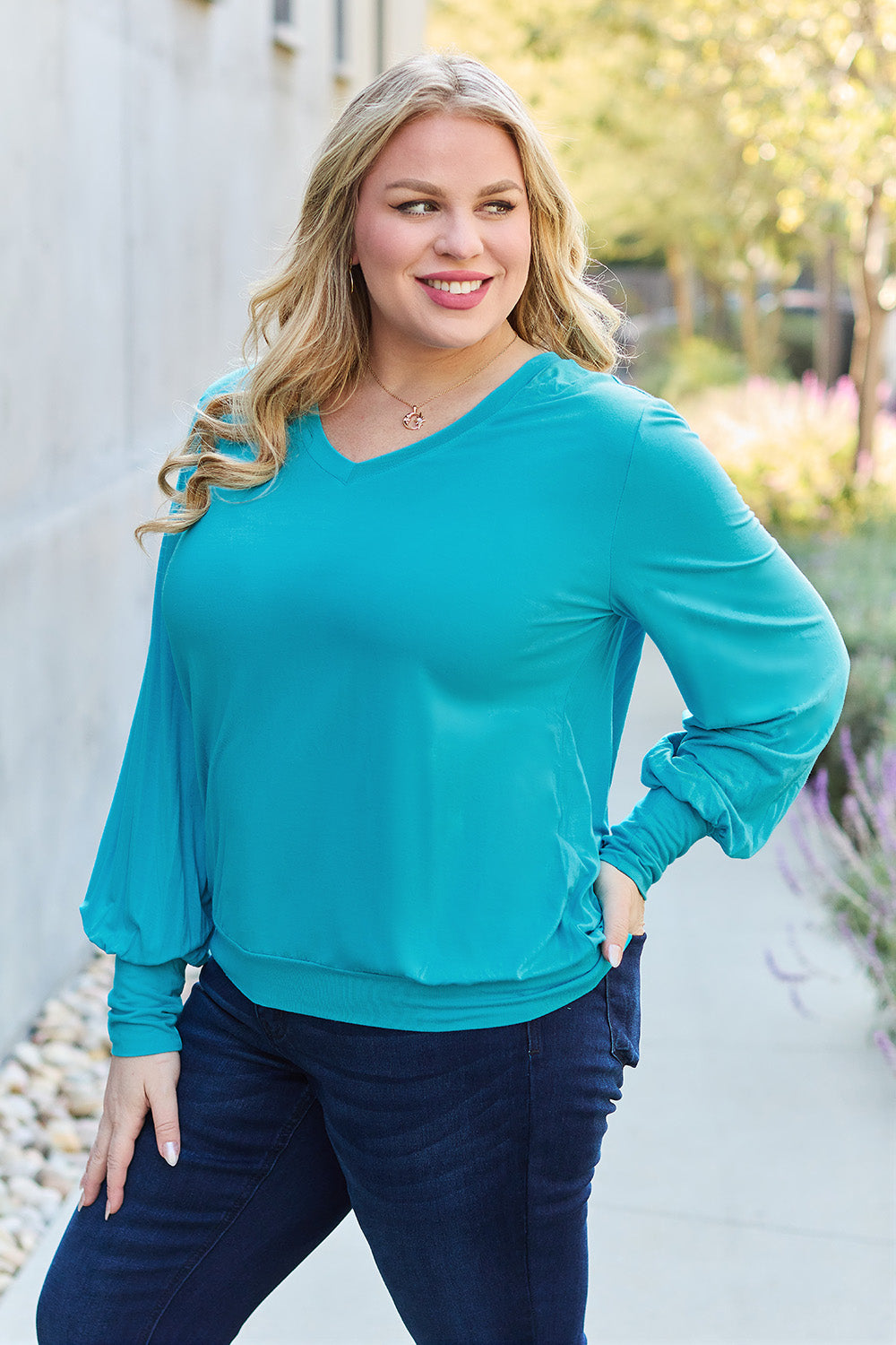 A woman with brown hair is standing against a concrete wall, wearing the Basic Bae Full Size V-Neck Lantern Sleeve Top in teal and blue basic-style jeans, smiling.