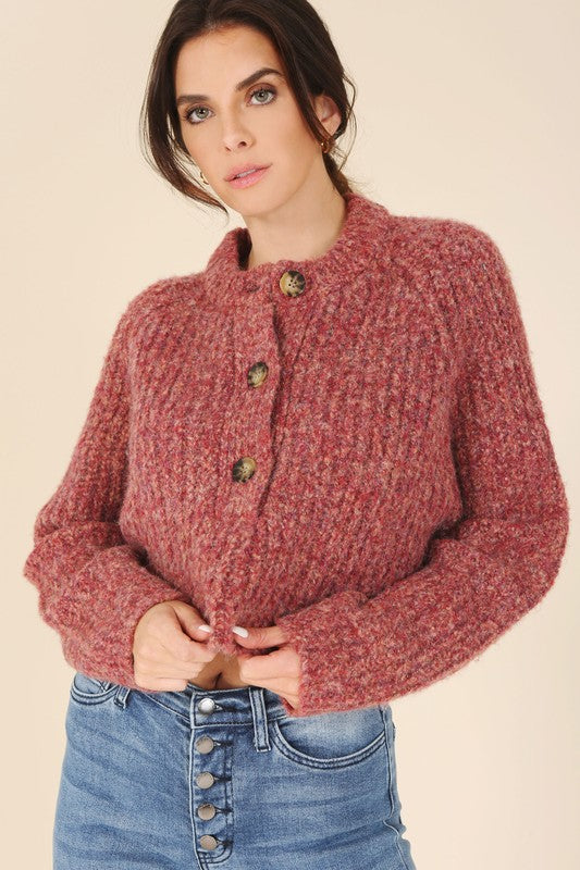 A person is wearing the Melange multicolor sweater top, characterized by textured pink fabric and featuring raglan sleeves along with large buttons, paired with light blue jeans, standing against a plain background.