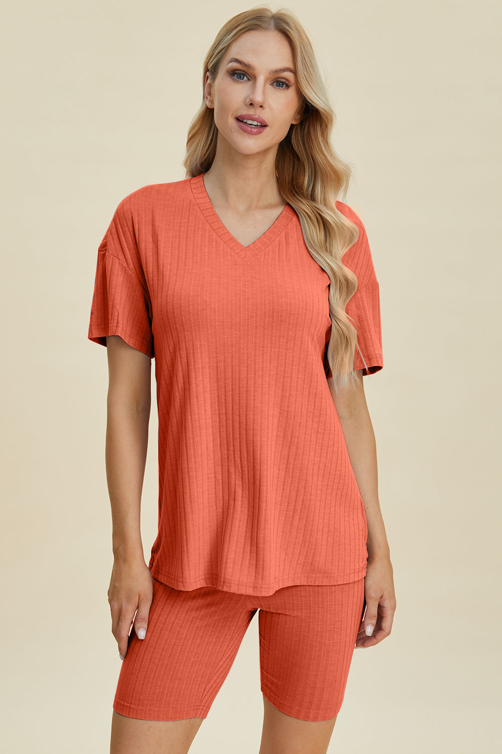 A woman with long blonde hair wearing the Basic Bae Full Size Ribbed V-Neck Short Sleeve Top and Shorts Set in rust stands against a plain background. This stylish two-piece set is made from a comfortable, stretchy fabric that is also machine washable.