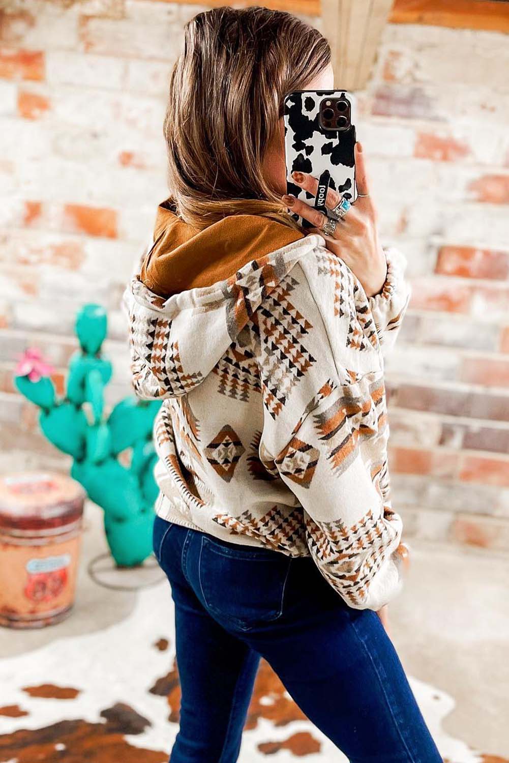 A person with long blonde hair, seen from behind, is wearing the Beige Aztec Print Kangaroo Pocket Half-Zip Hoodie. The hoodie showcases an Aztec pattern in shades of brown, beige, and orange and features a drop shoulder design with a cozy kangaroo pocket, all while standing outdoors.