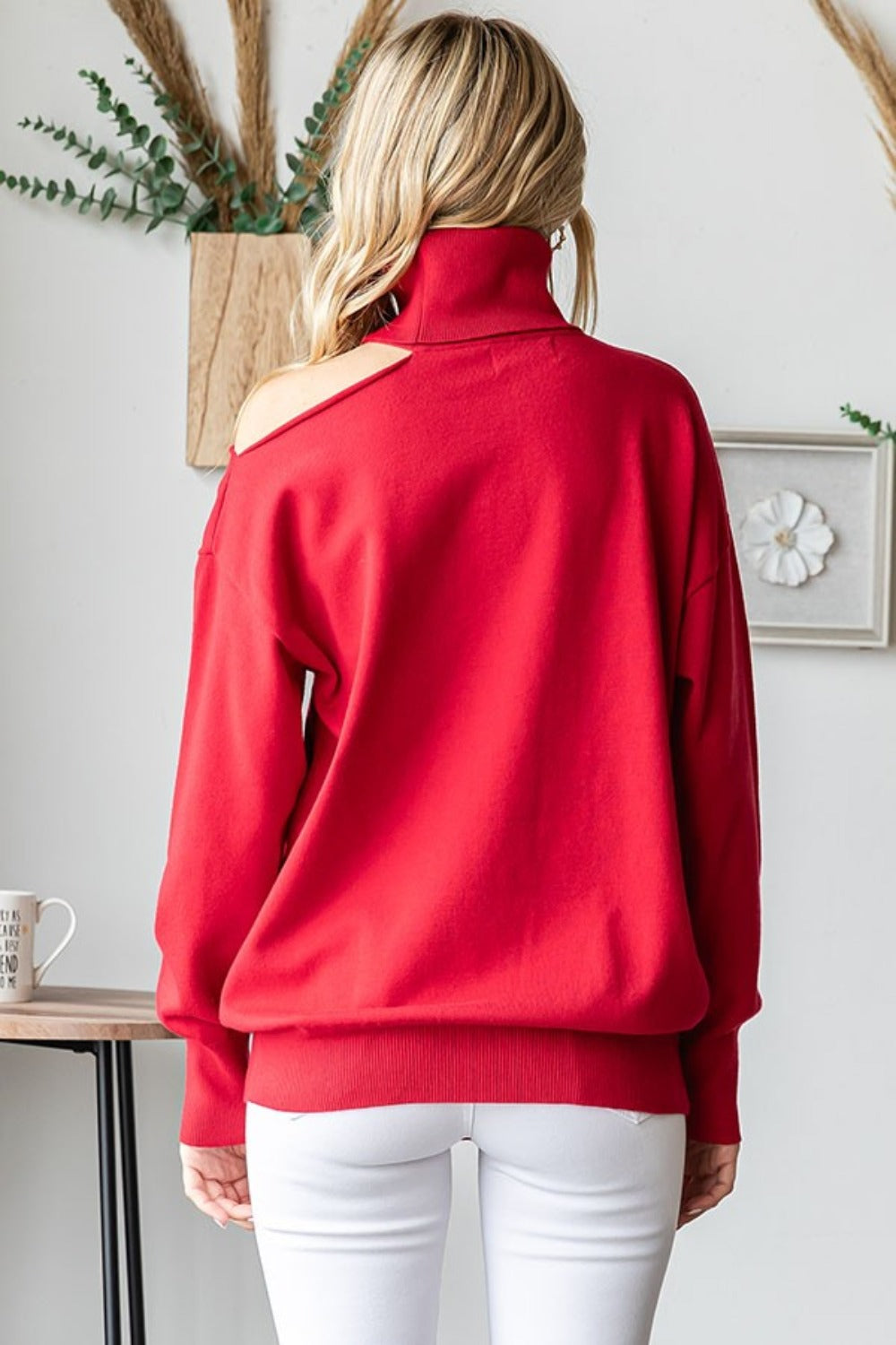 A blonde woman in the stylish First Love Cut Out Shoulder Turtleneck Sweater and white pants sits on a stool against a wall, with plants in the background.