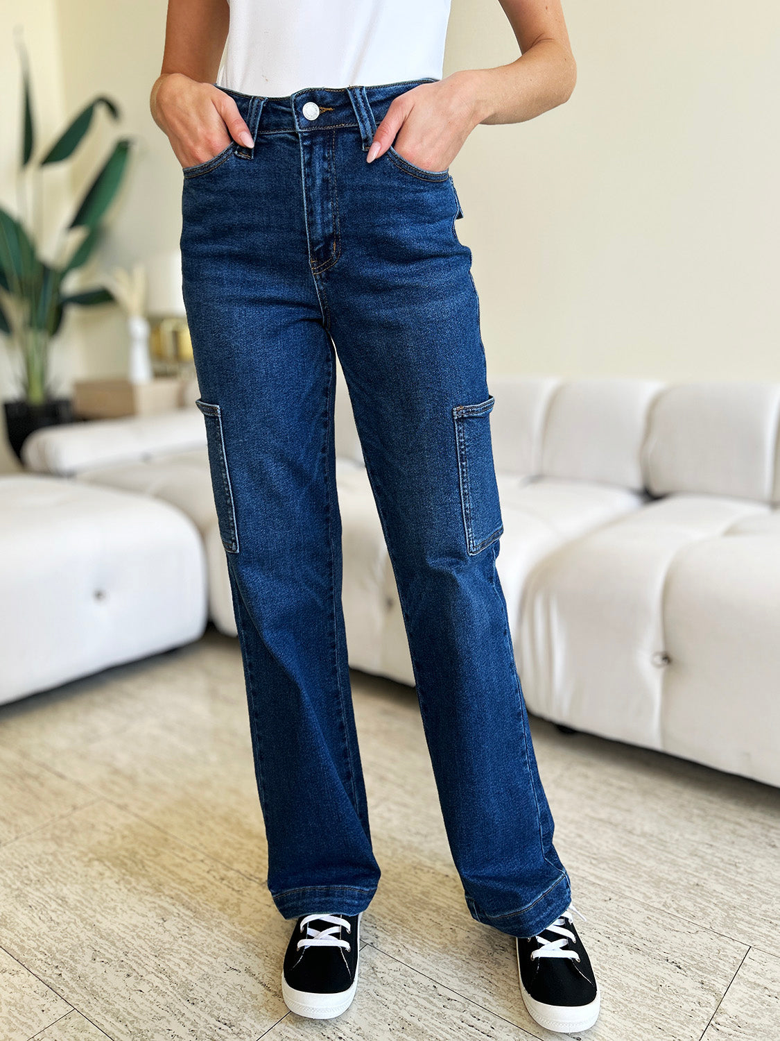 A person wearing Judy Blue Full Size High Waist Straight Cargo Jeans made of durable denim fabric and a white top walks on a wooden floor, with a white sofa and green plant in the background.