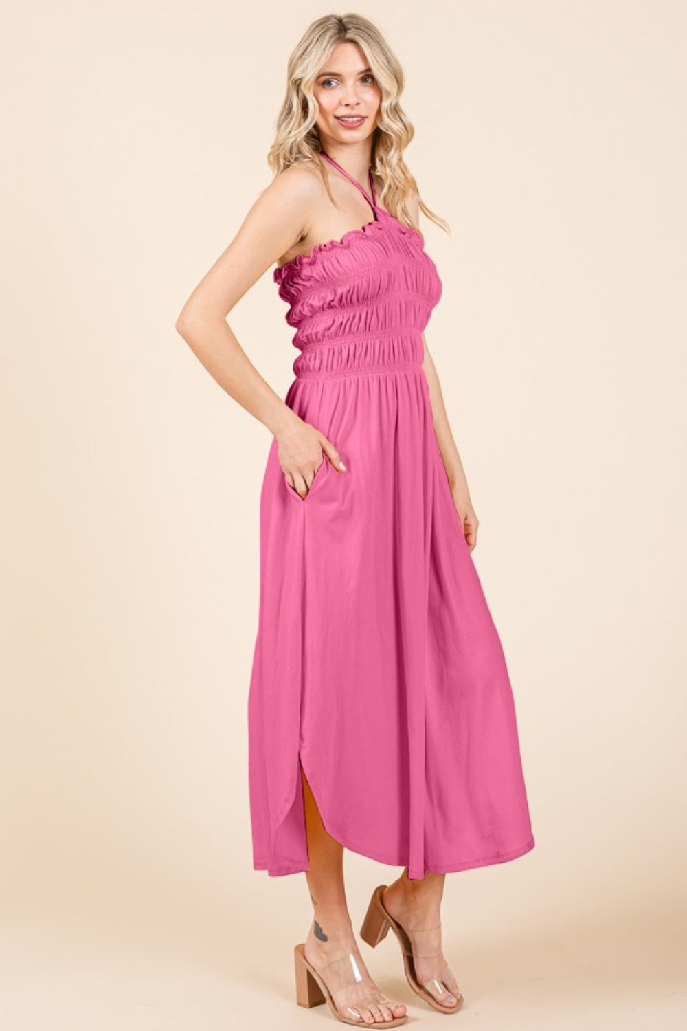 Someone wearing a Culture Code Tie Back Shirring Dress with pockets, featuring a sleeveless and smocked pink halter style, standing against a beige background.