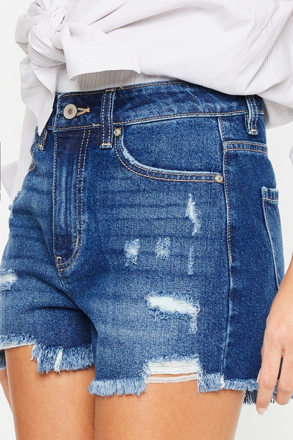 A woman with long dark hair wears a white blouse tied at the waist over a white top and Kancan High Rise Frayed Hem Denim Shorts, posing with one hand in her pocket against a plain background, embodying the perfect summer wardrobe.