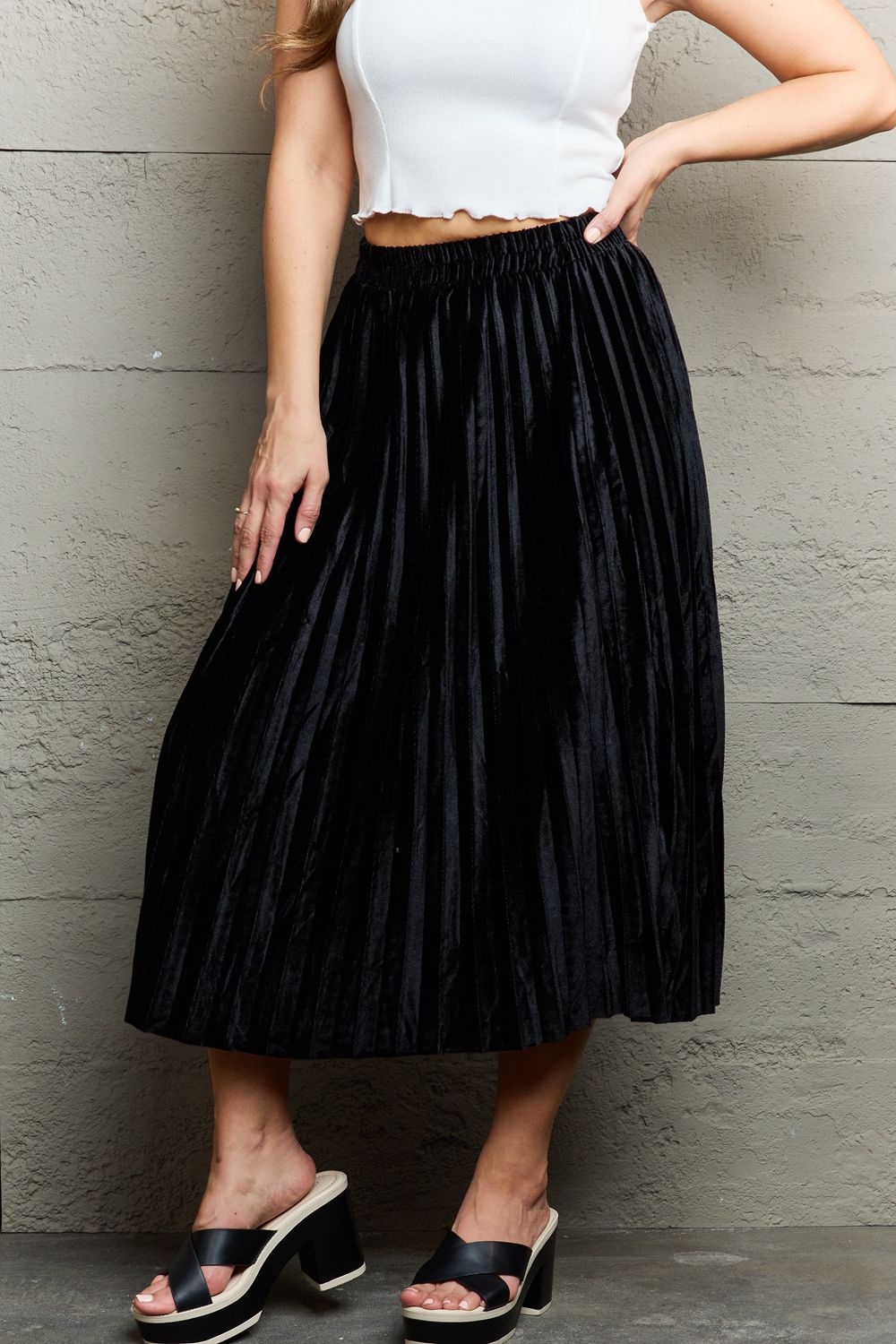 Person wearing a white sleeveless top, an elegant Ninexis Accordion Pleated Flowy Midi Skirt, and black platform sandals stands against a textured gray wall.