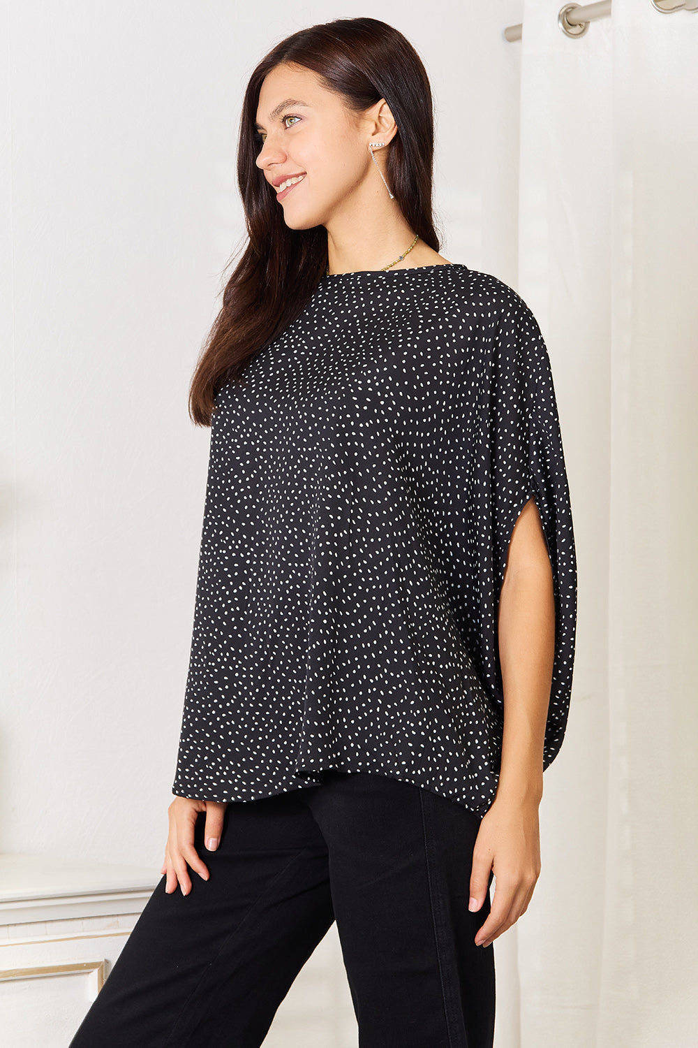 A woman stands wearing the Perfee Printed Dolman Sleeve Round Neck Blouse with white dots and black pants, smiling and looking to her left. The blouse features a printed design that adds a touch of elegance to her outfit.