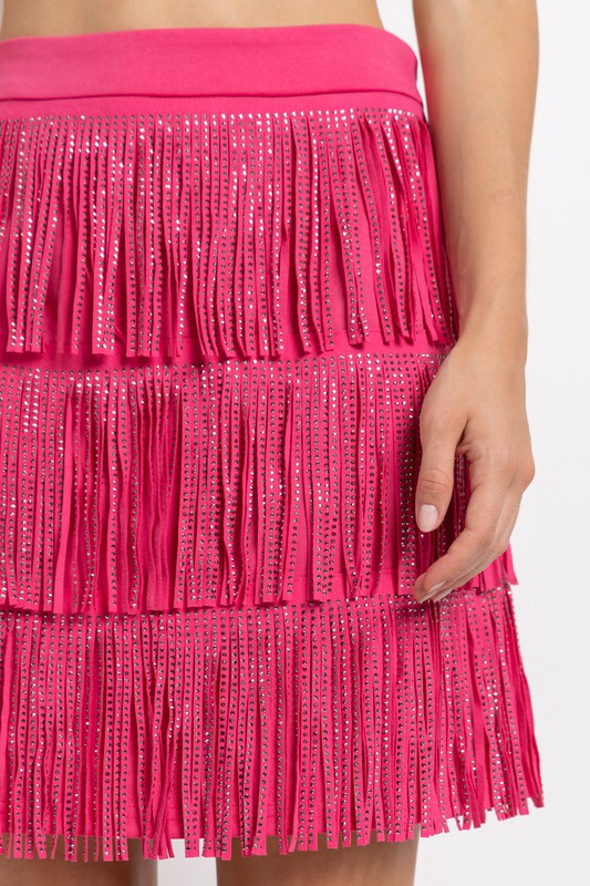 Close-up of the RHINESTONE SUEDE FRINGE SKIRT in pink, featuring a person’s hand resting lightly on the skirt adorned with rhinestone fringes.