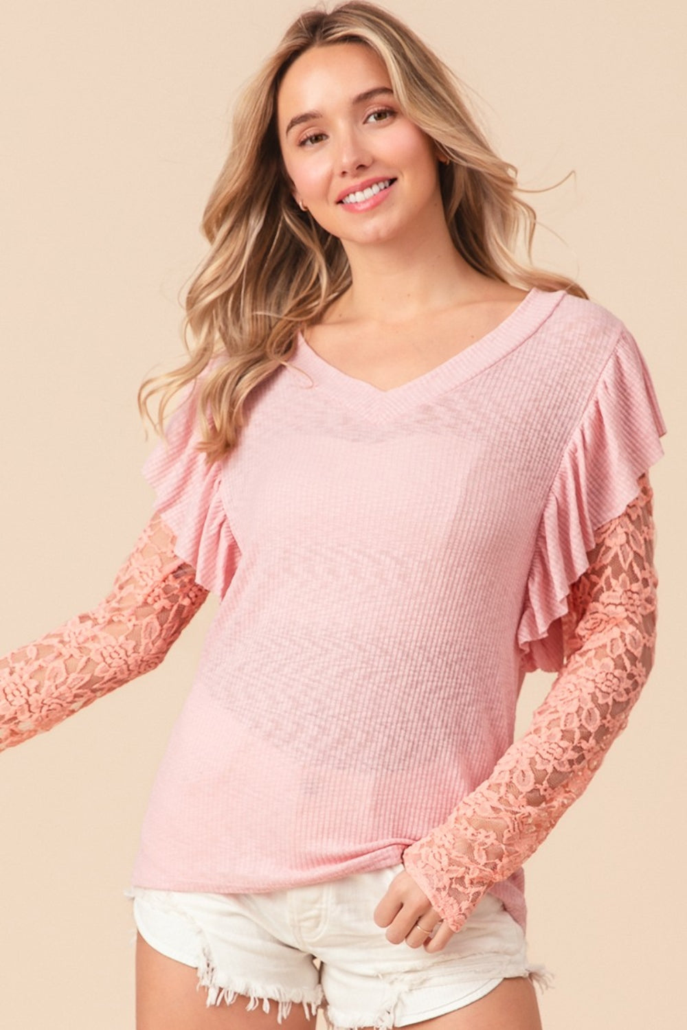 A woman with long blonde hair, smiling and looking to the side, is wearing a pink BiBi Ruffled Lace Sleeve Rib Knit Top paired with white shorts.