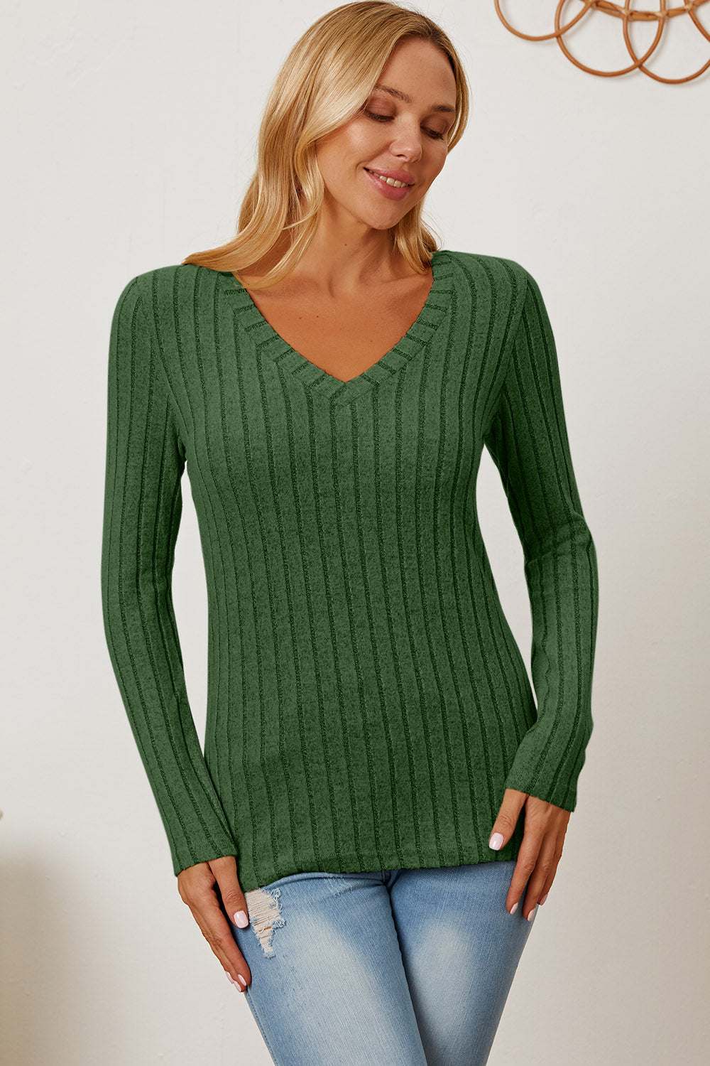 A blonde-haired woman smiles while wearing the Basic Bae Full Size Ribbed V-Neck Long Sleeve T-Shirt, which is light green and slightly stretchy, paired with light blue jeans. She holds one hand near her head and the other near her waist.