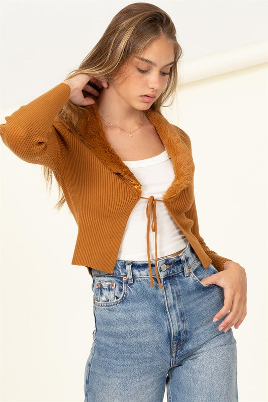 A person stands wearing the Miss Mesmerize Fur Trim Tie Front Ribbed Cardigan in brown, a white top, and blue jeans, with their right hand in their pocket.