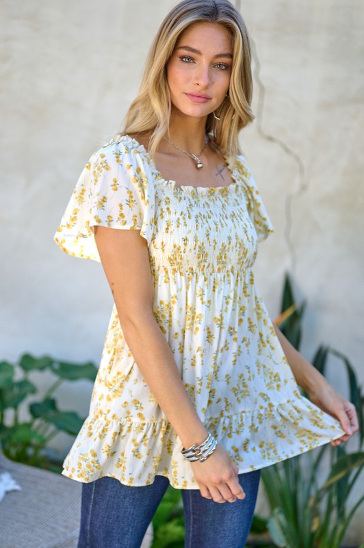 A woman stands outdoors donning a Floral Printed V-Neck Ruffle Top paired with jeans, set against a neutral background, with plants in the foreground, highlighting her fashion-forward wardrobe.