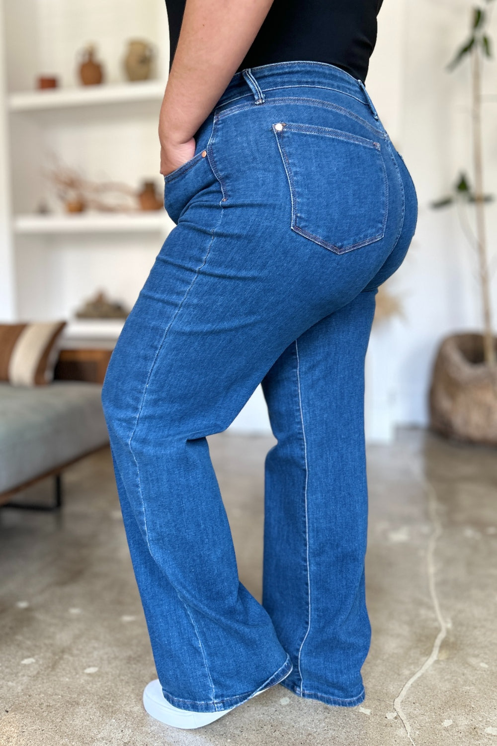 A person wearing Judy Blue Full Size High Rise Straight Jeans, a black top, and white shoes stands gracefully on a polished concrete floor.