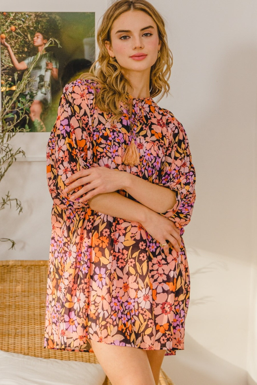 A woman in a flattering silhouette stands indoors with arms folded, wearing the vibrant ODDI Full Size Floral Tied Neck Mini Dress. She poses gracefully in front of a framed picture and wicker chair.