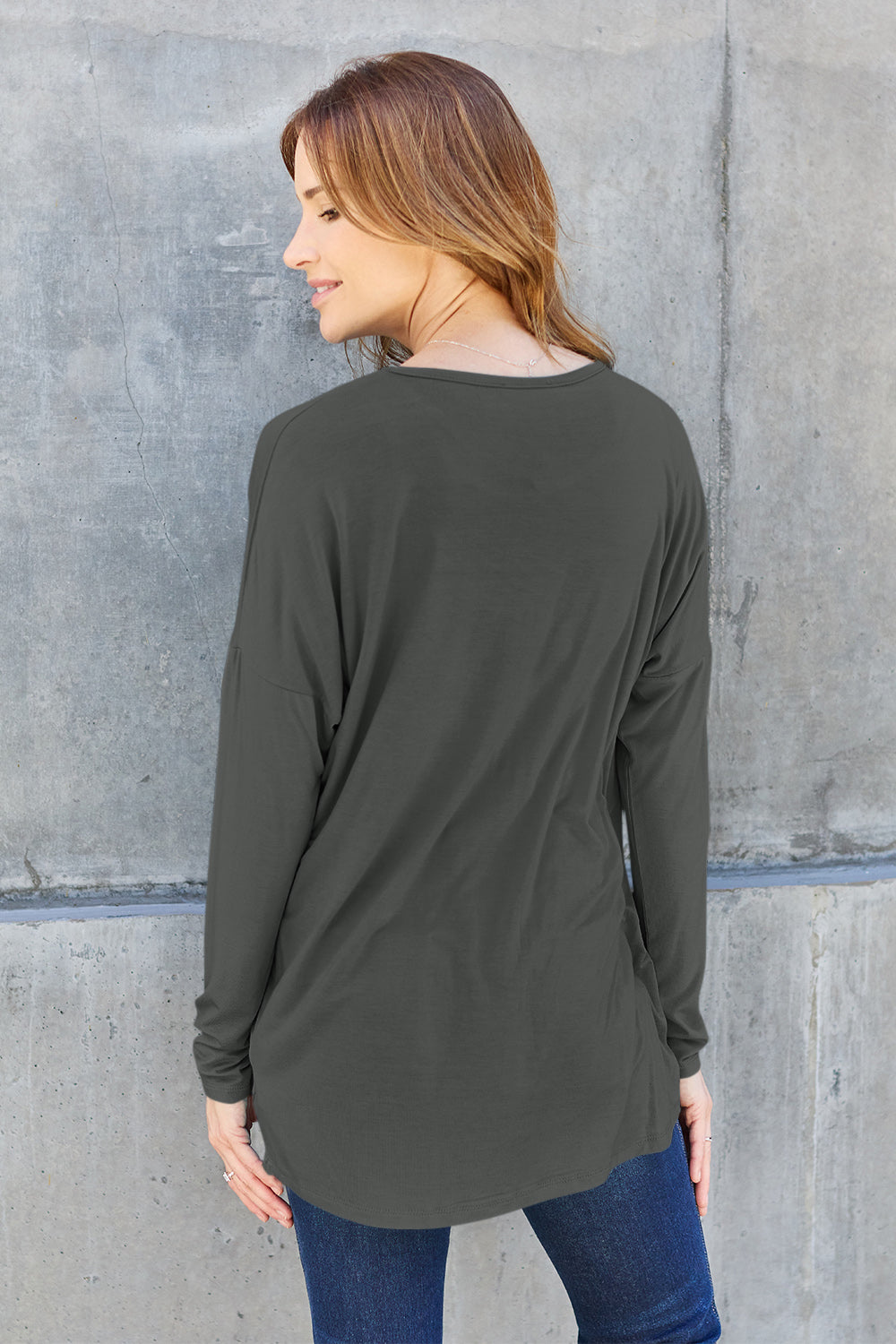 A woman with shoulder-length hair wearing a Basic Bae Full Size Round Neck Dropped Shoulder T-Shirt in green and a pair of jeans, exuding a basic style, stands against a concrete wall. She's holding an imported brown woven backpack on her shoulder while touching her hair with her hand.