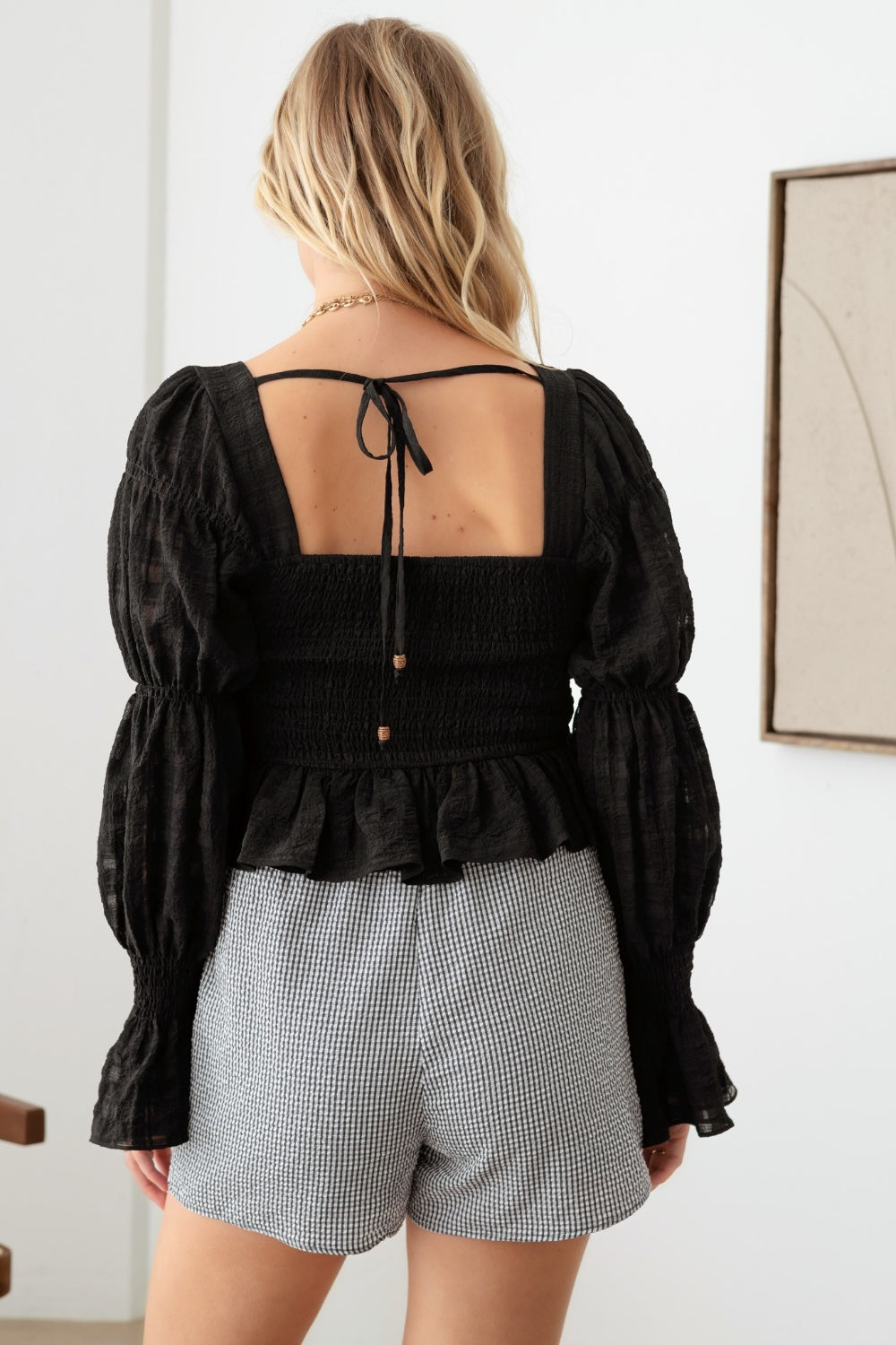 A person with long blonde hair is standing indoors, wearing a Mustard Seed Shirred Smocked Peplum Long Sleeve Top and checkered shorts for a trendy feminine look. They are gazing directly at the camera with hands resting lightly on their hips.