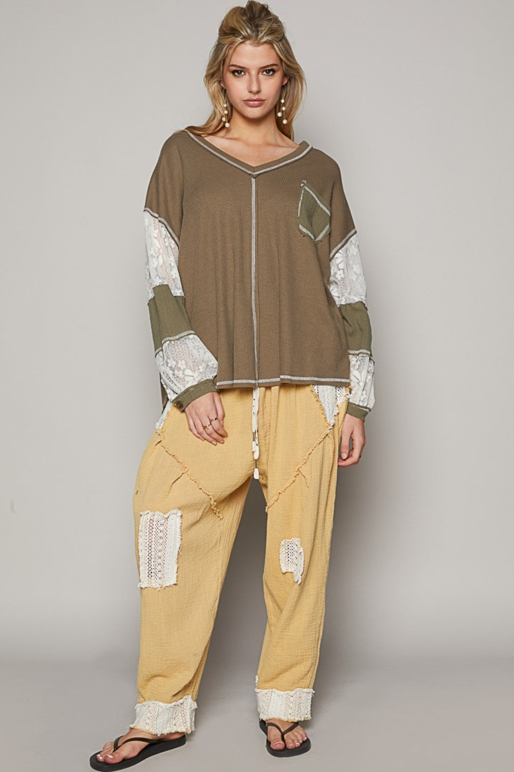 A person stands against a plain background, wearing a loose-fitting, patchwork outfit in shades of brown and beige. The ensemble features the POL V-Neck Lace Balloon Sleeve Exposed Seam Top paired with matching pants. They complete the look with black sandals.