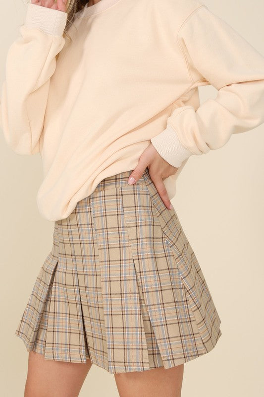 A person dressed in a plaid pleated mini skirt paired with a beige sweater is posed against a plain backdrop. This fashionable and versatile outfit, featuring the Plaid Pleated Mini Skirt, is designed for easy maintenance and can be machine washed in cold water.