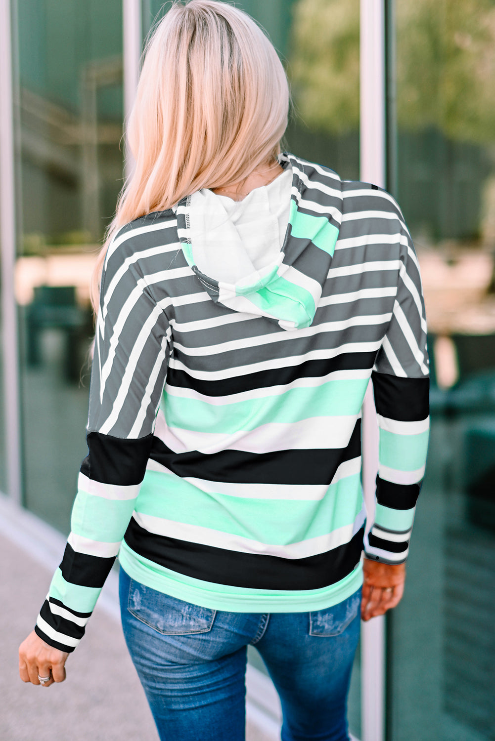 A person with long blonde hair, dressed in a green striped drawstring long sleeve hoodie and jeans, stands in front of a building with glass windows, facing away from the camera.