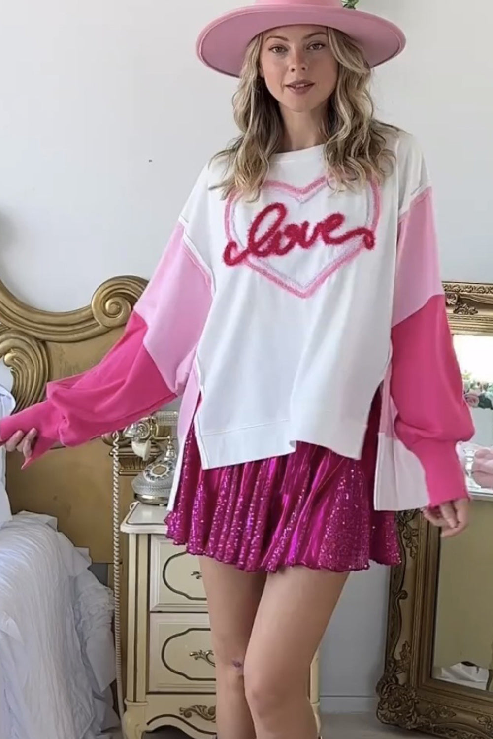 A woman with long blonde hair is smiling and posing indoors. She is wearing a White Tinsel Love Heart Graphic Color Block Sleeve Sweatshirt with pink shorts. There are framed photos and a table in the background.