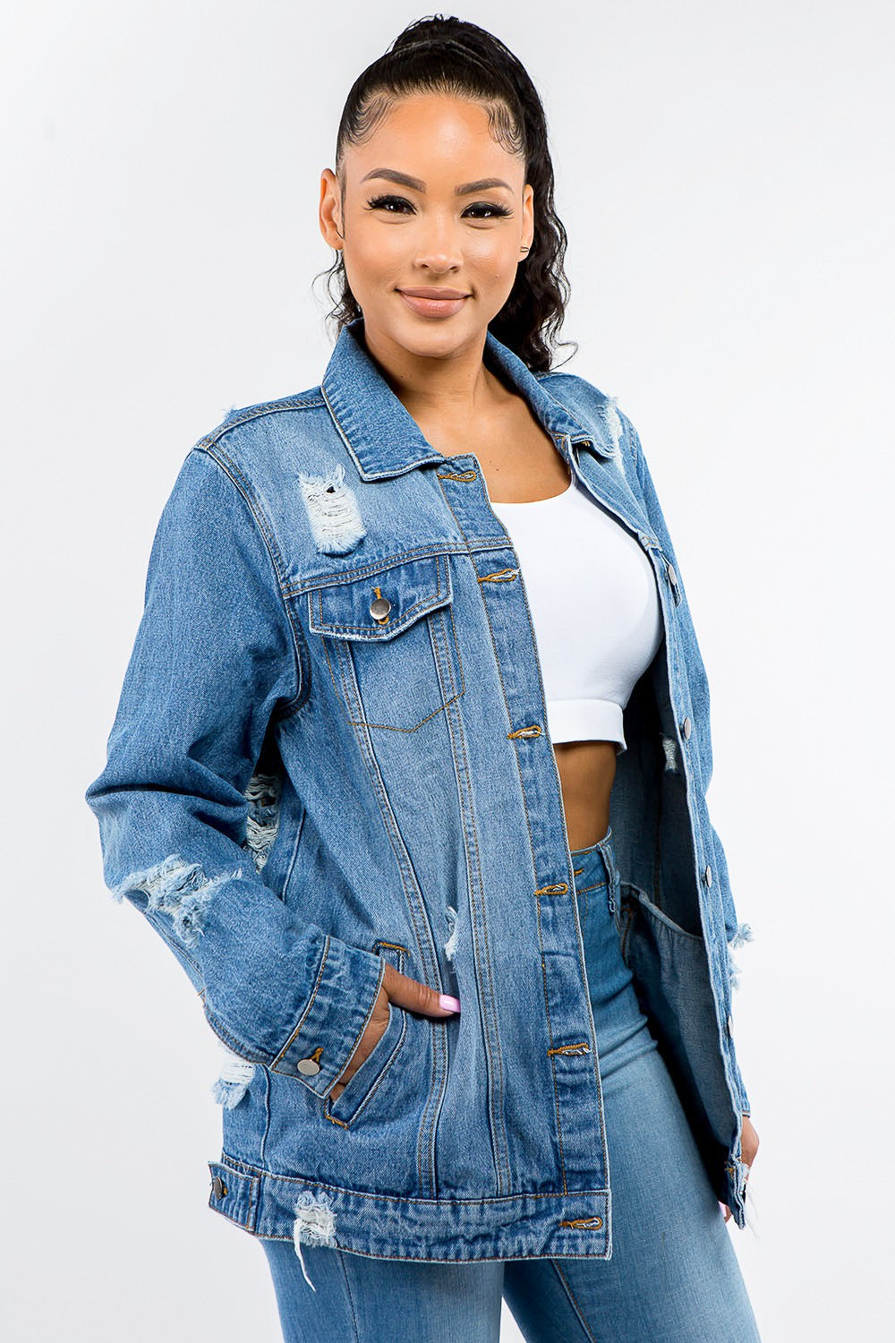 A woman in an American Bazi Full Size Button Up Distressed Denim Jacket with a vintage look stands against a plain background, pairing it perfectly with a white crop top and denim jeans.