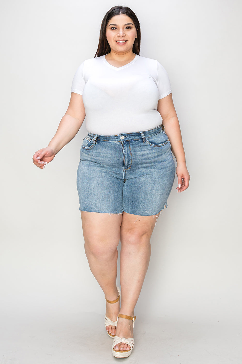 Side view of a person wearing a white shirt and Judy Blue Full Size High Waist Raw Hem Denim Shorts, standing indoors near a wooden cabinet. Summer fashion vibes radiate through the casual ensemble.