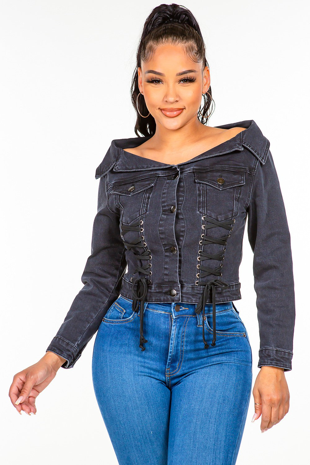 A woman in an American Bazi Off Shoulder Lace Up Denim Jacket and blue jeans stands against a plain white background. She has long hair tied up in a high ponytail.