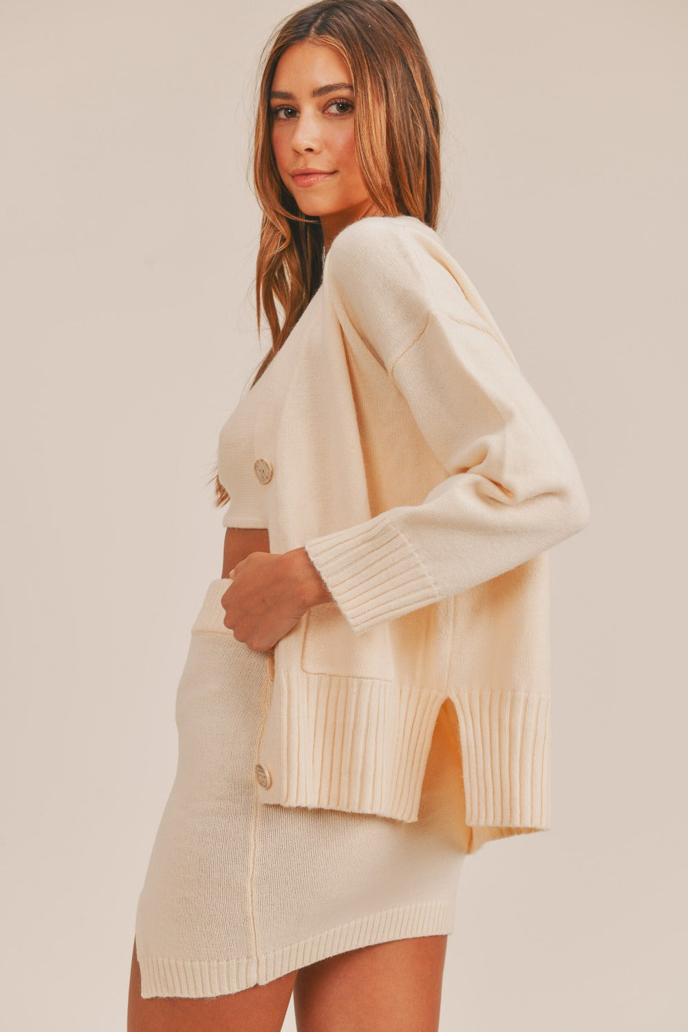 A woman wears the MABLE 3 Pieces Sweater Set, featuring a cream-colored cardigan over a matching crop cami and high-waisted mini slit skirt. She has long hair and is looking slightly off to the side.