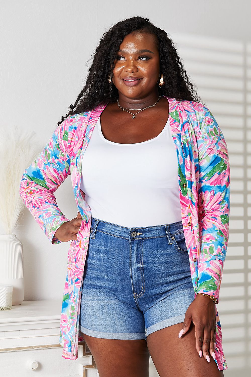 Wearing the Double Take Floral Open Front Long Sleeve Cardigan in shades of pink and blue over a white top and denim shorts, a person exudes feminine charm while standing in a well-lit room, smiling.