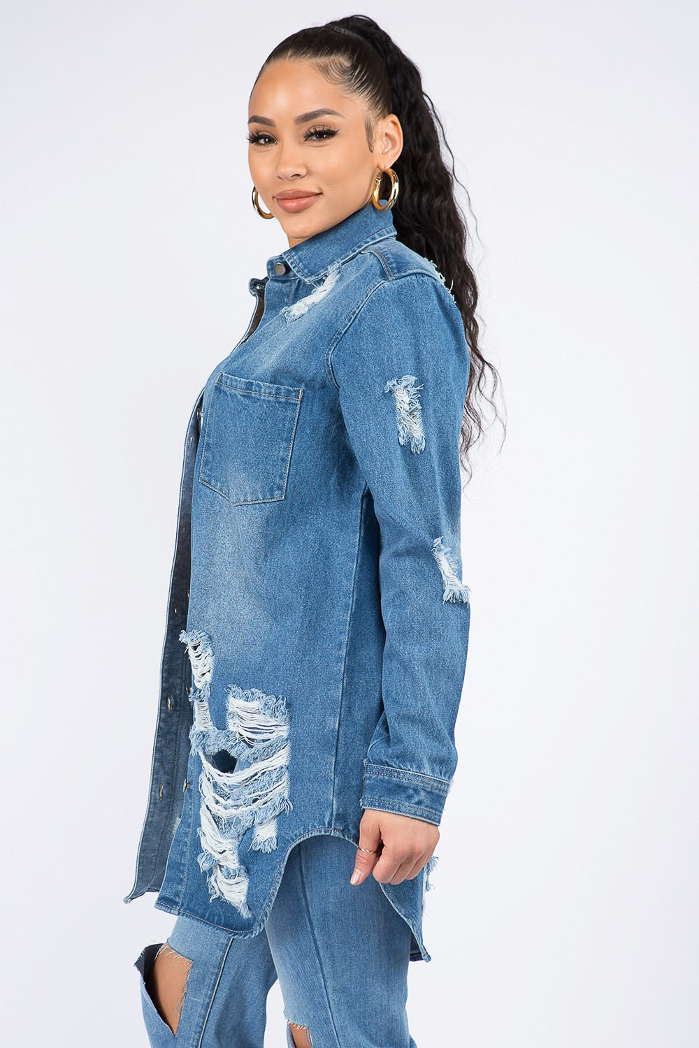 A woman wearing the American Bazi Distressed Button Down Denim Shirt Jacket and jeans paired with a white crop top exudes vintage-inspired charm as she poses with one hand on her head against a plain background.