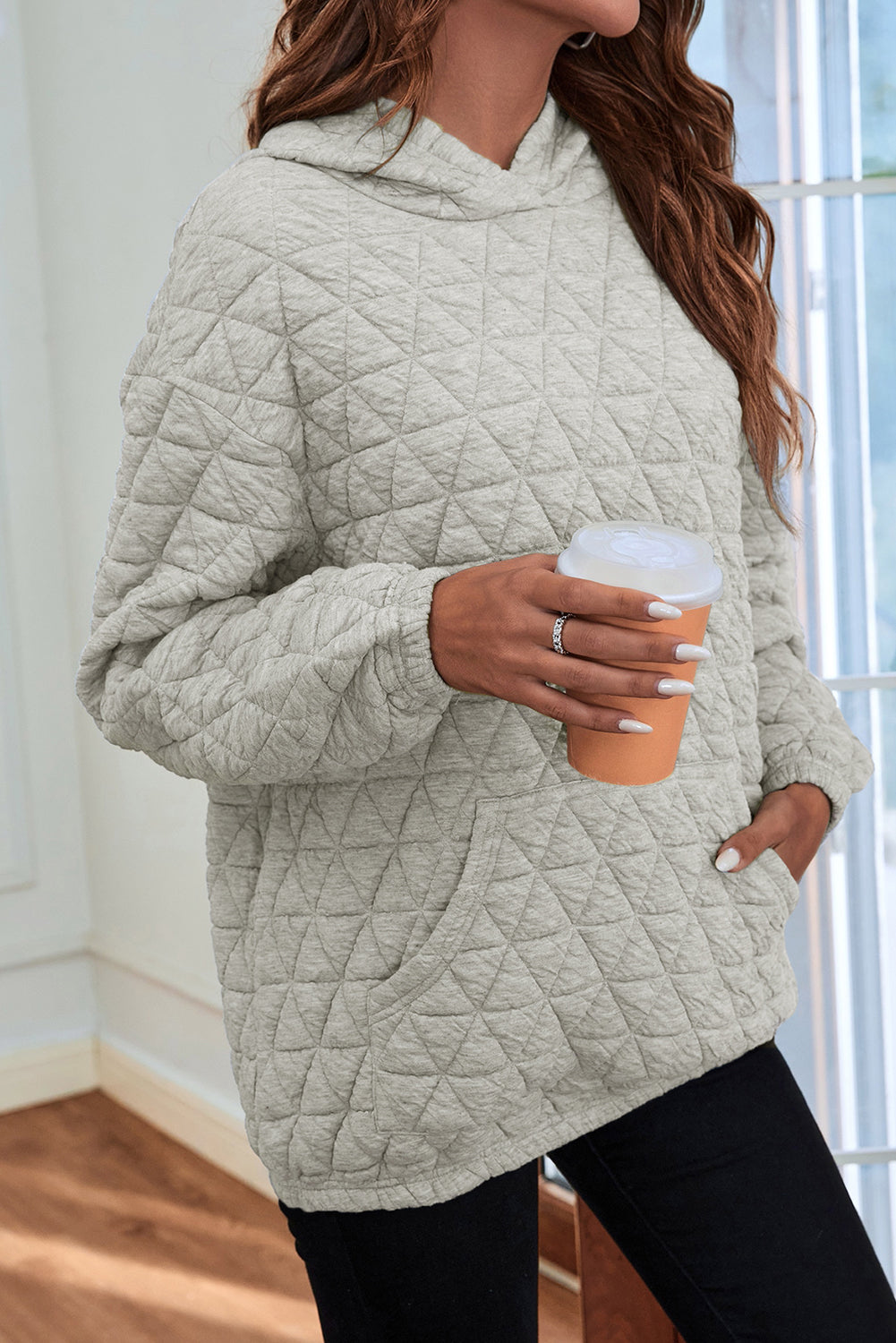 A woman stands partially turned away, wearing a Light Grey Solid Color Quilted Kangaroo Pocket Hoodie with a drop shoulder design and blue jeans. She touches her hair with her right hand, and there are beige and brown decor elements in the background.