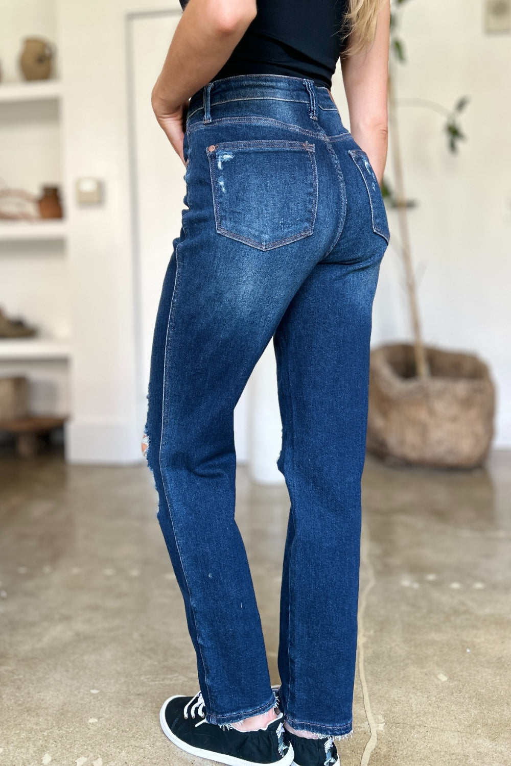 A woman in a black sleeveless top and Judy Blue Full Size High Waist Rigid Magic Heavy Destroy Straight Jeans stands indoors with her hands in her pockets. The background features a plant and shelves adorned with decor items.