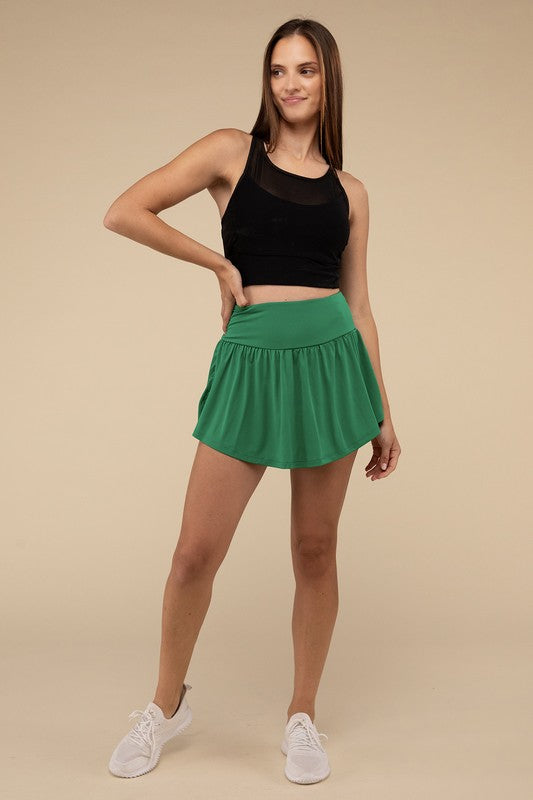 Close-up of a woman in a black crop top and Wide Band Tennis Skirt with Zippered Back Pocket, pulling the skirt to show matching shorts with a zippered back pocket underneath.