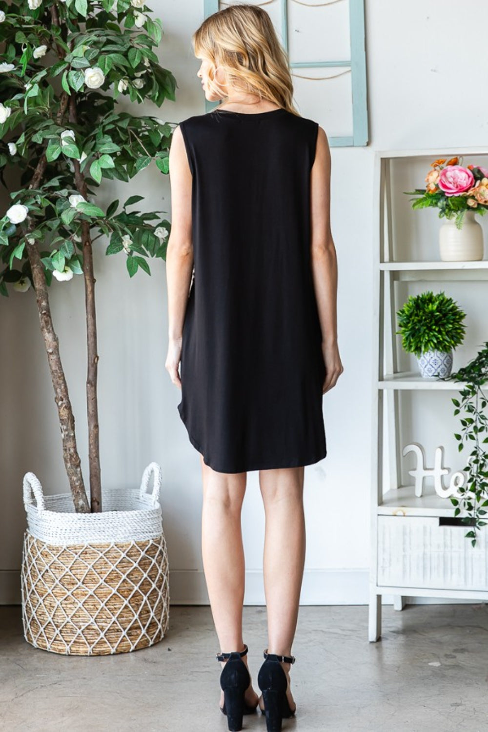 A woman wearing the Heimish Full Size Sleeveless Animal Print Mini Dress and black heels stands indoors beside a potted plant and a shelf adorned with decor items, creating a striking impression.