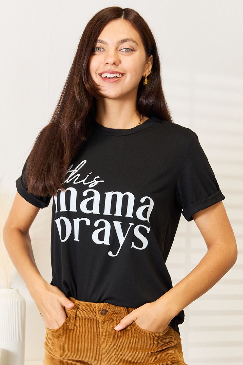 A woman wearing a Simply Love THIS MAMA PRAYS Graphic T-Shirt, paired with brown pants, stands gracefully in a softly lit room, exuding a sense of calm and spiritual gift.