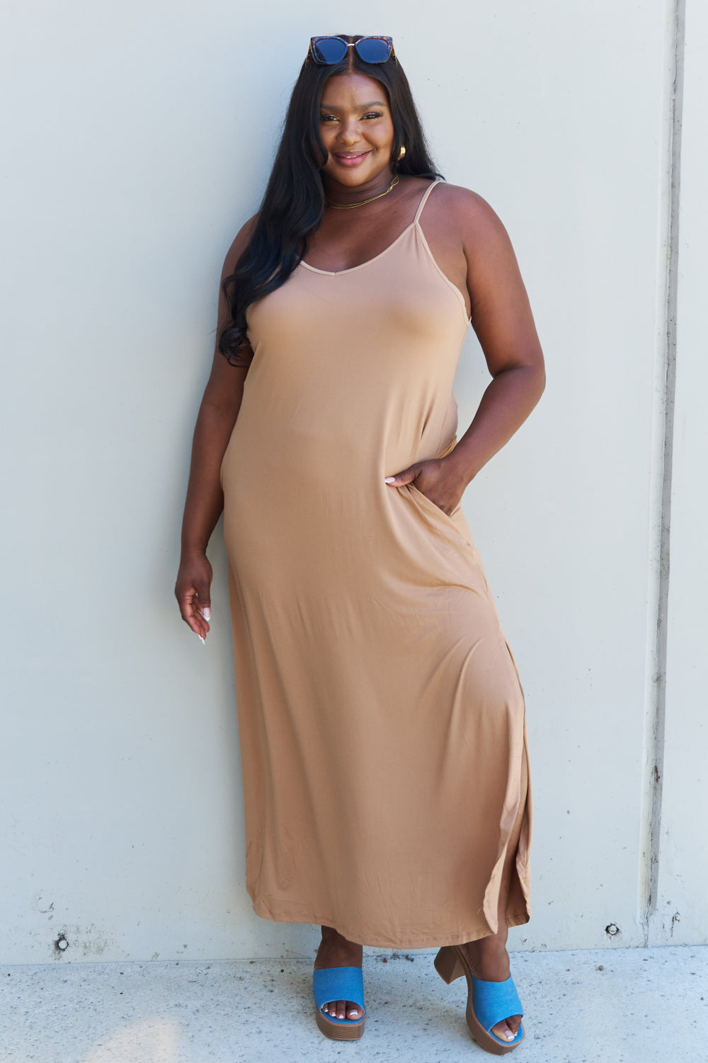 A woman stands against a plain background, wearing the Ninexis Good Energy Full Size Cami Side Slit Maxi Dress in Camel along with a wide-brimmed hat. She has one hand on her hip and is smiling.