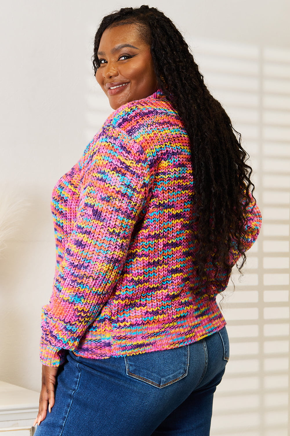 Person wearing an Angel Wings Woven Right V-Neck Long Sleeve Cardigan, characterized by its multicolored design and large buttons, paired with blue jeans. The cardigan, made of 100% acrylic knit, offers a versatile look perfect for any occasion. They are standing indoors against a white background.