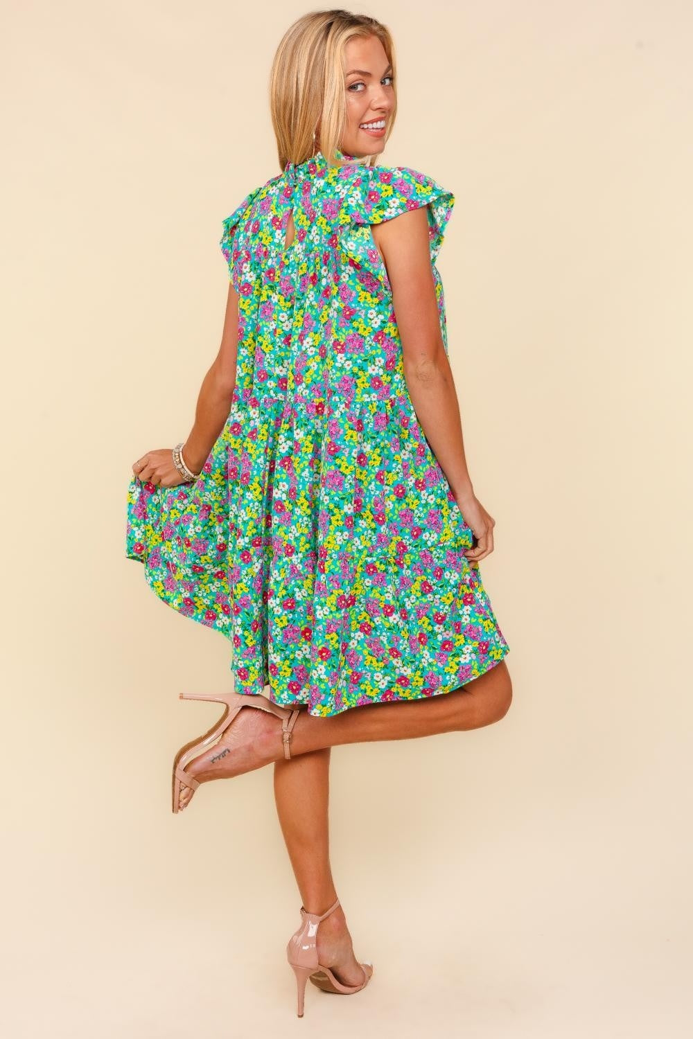 A woman in a Haptics Frilled Mock Neck Ditsy Floral Dress stands against a plain background, her beige high heels accentuating the feminine, elegant silhouette. Her hands rest casually in the pockets of her colorful, ditzy small floral print dress with flutter short sleeves.