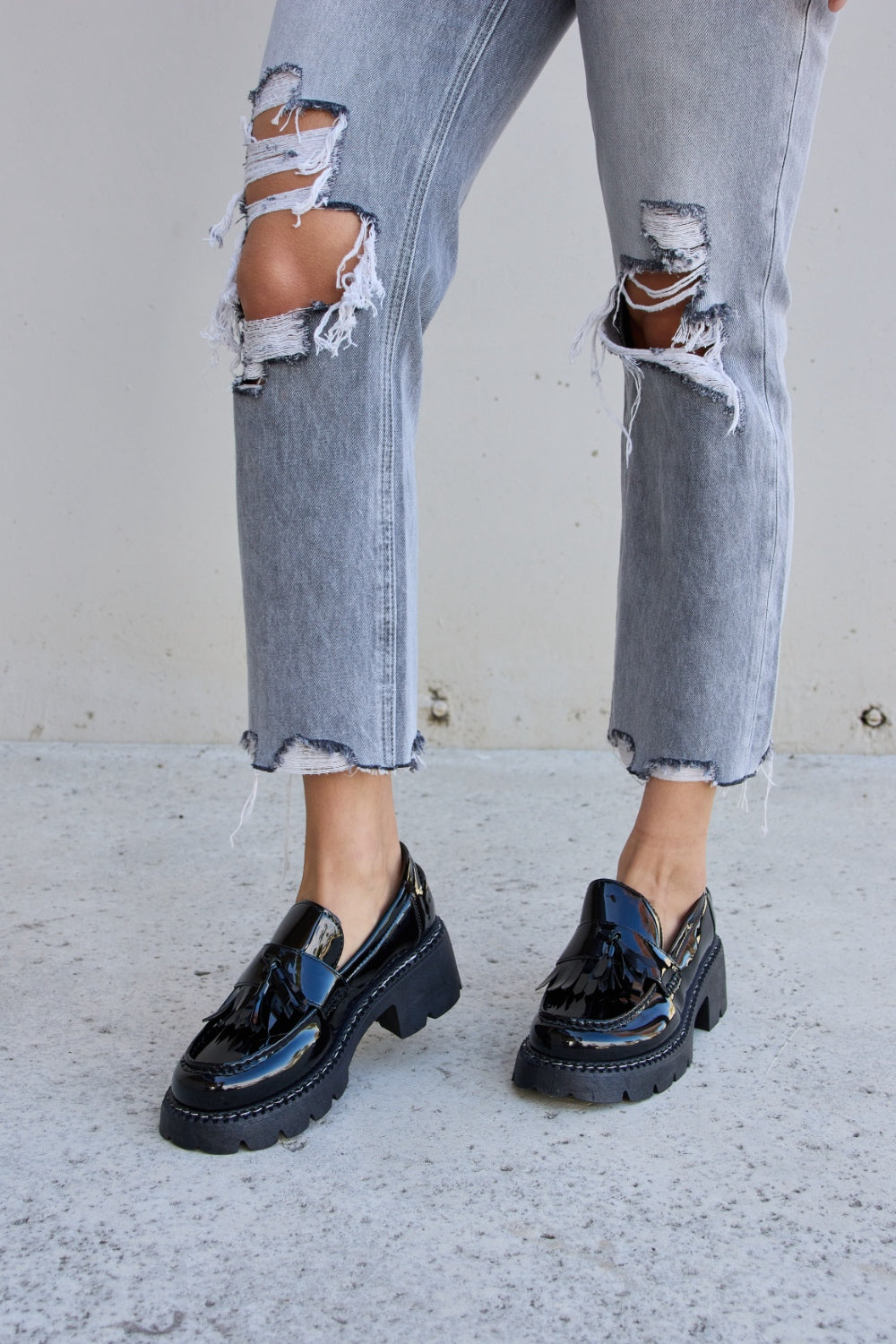 A person wearing ripped gray jeans and the Forever Link Tassel Detail Mid Heel Chunky Loafers, which feature shiny black tones, chunky soles, and a subtle tassel detail.