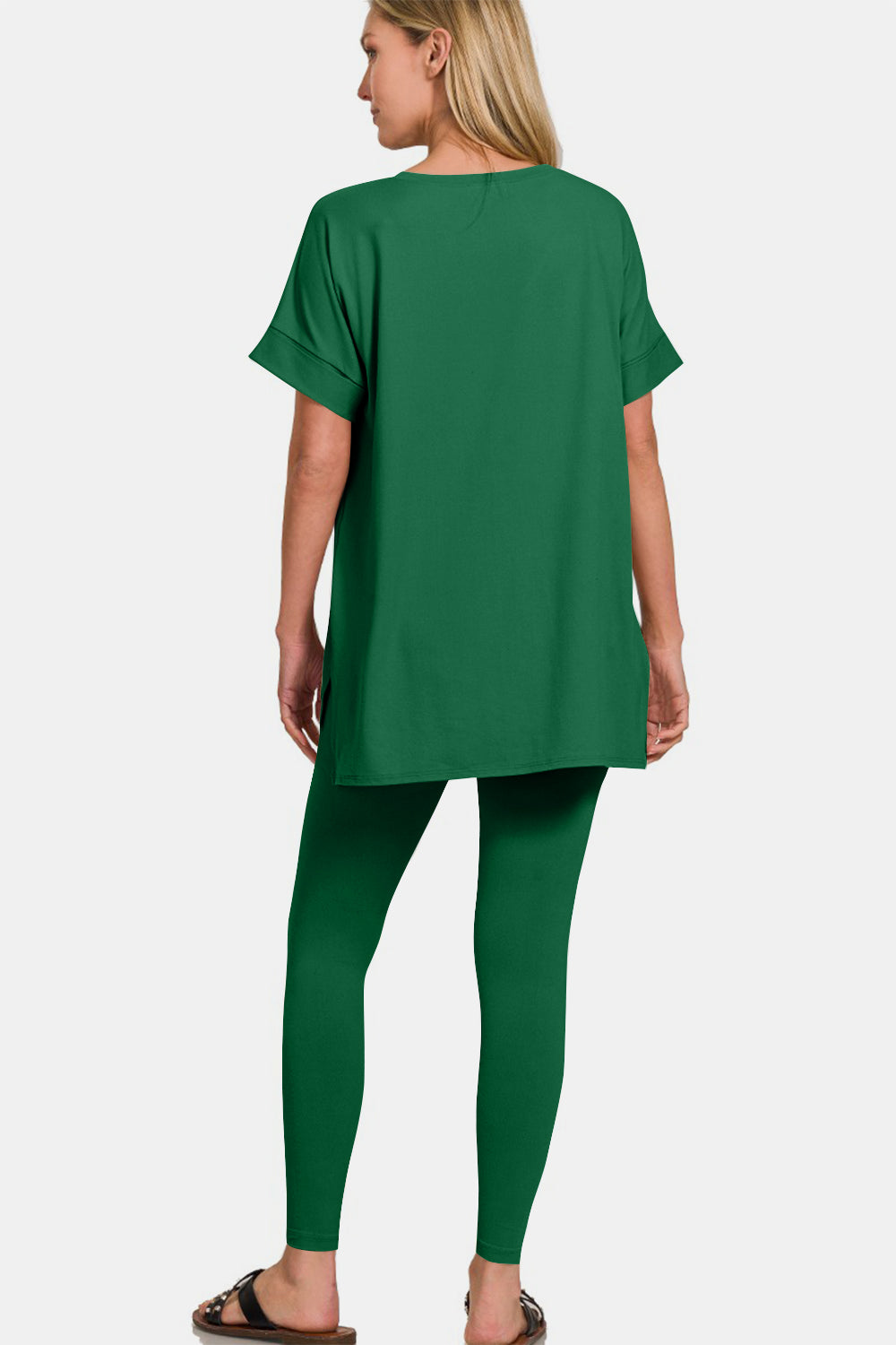 A person wearing the Zenana V-Neck Rolled Short Sleeve T-Shirt and Leggings Lounge Set in green, standing and smiling with one hand touching the side of their head, stylishly matched with sandals.