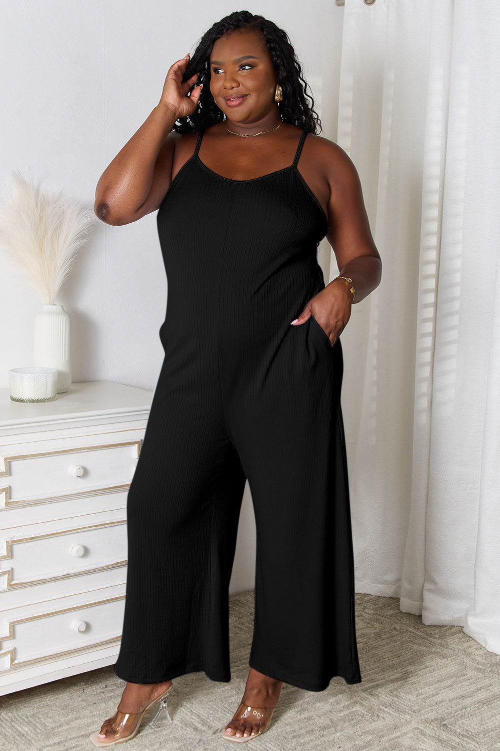 A woman in a stylish Basic Bae Full Size Spaghetti Strap V-Neck Jumpsuit stands indoors near a white dresser adorned with decorative items, smiling with one hand in her pocket.