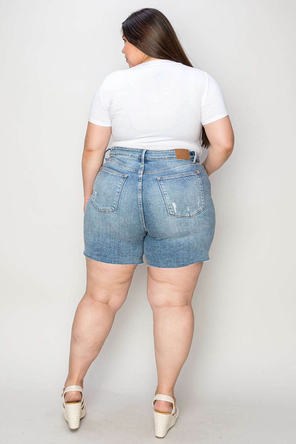 A woman wearing a white T-shirt, Judy Blue Full Size High Waist Rhinestone Decor Denim Shorts, and white sandals poses against a plain background.