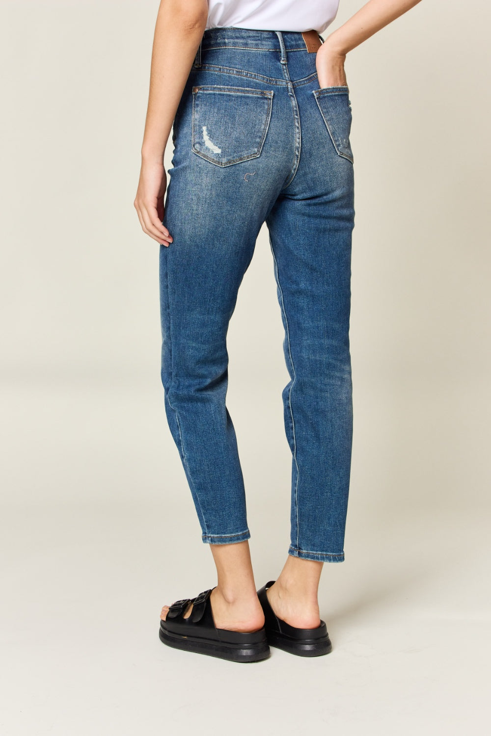 Person wearing Judy Blue Full Size Tummy Control High Waist Slim Jeans in blue and a white shirt, with their hands in their pockets. They are standing indoors on a polished concrete floor with a wooden cabinet and plant in the background.