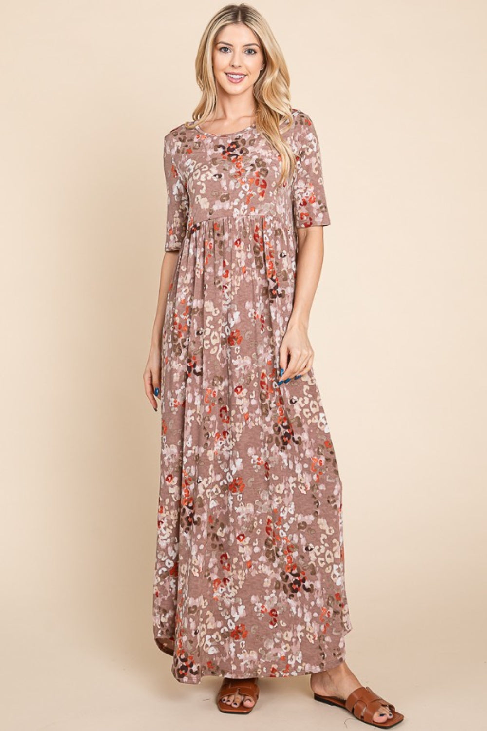 A woman stands against a beige background wearing the light brown, floral BOMBOM Printed Shirred Maxi Dress and brown sandals. She has long blond hair and is looking to her right with one hand slightly raised. The versatile dress offers a flattering fit perfect for any occasion.