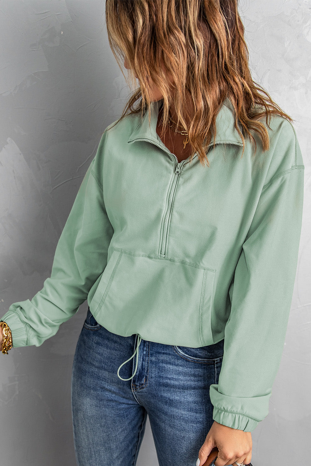 A person with wavy hair stands with their back to the camera, wearing a Green Zip Front Pocketed Pullover Sweatshirt and blue jeans, holding a small brown bag in their left hand.