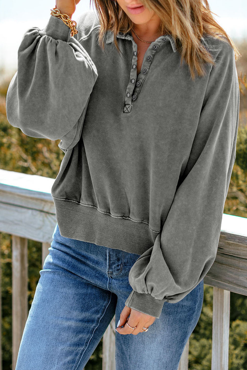 A person with long hair stands outdoors, facing away, wearing a chic **Gray Washed Snap Buttons Lantern Sleeve Pullover Sweatshirt** and blue jeans, resting their hands on a wooden railing.