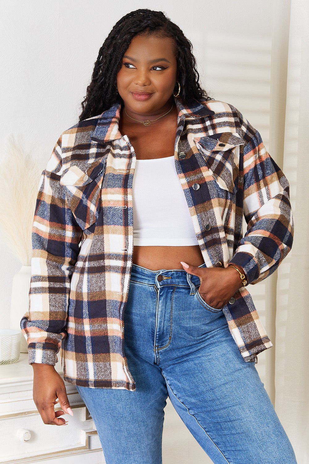 A person with long braided hair wearing the Double Take Plaid Button Front Shirt Jacket with Breast Pockets over a white top and blue jeans stands with a hand in their pocket, posing in an indoor setting. The layering of the jacket adds an effortless touch to the outfit.