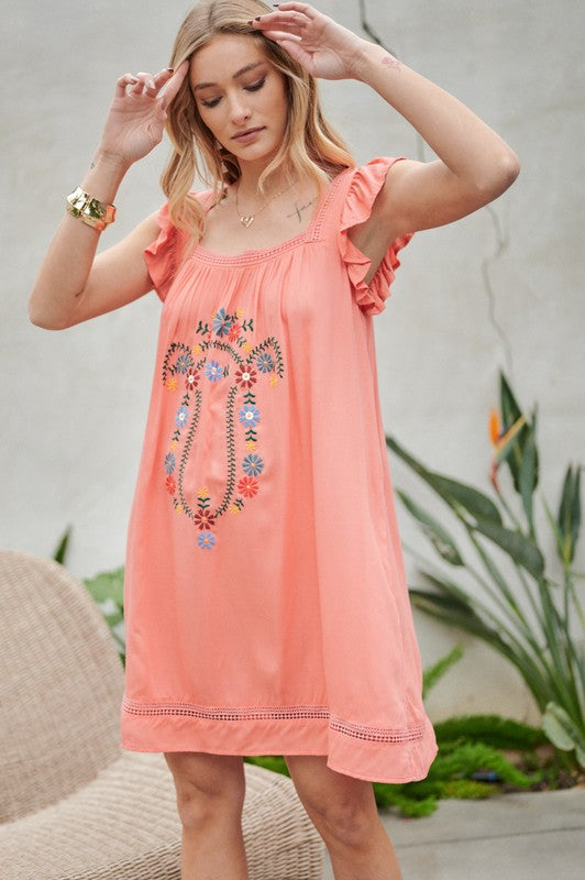 A woman in an Embroidered Square Neck Mini Dress, featuring a coral hue, stands near plants adjusting her hair—the perfect summer attire.