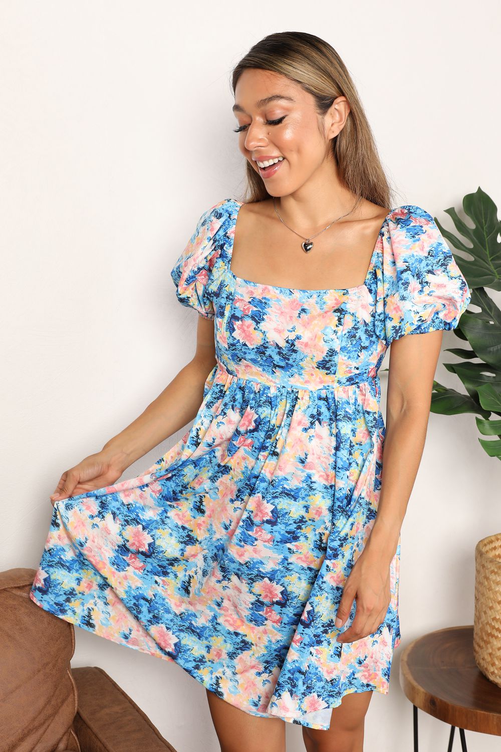 A woman in the Honey Floral Square Neck Puff Sleeve Dress stands smiling indoors, with a plant and a brown chair visible in the background.