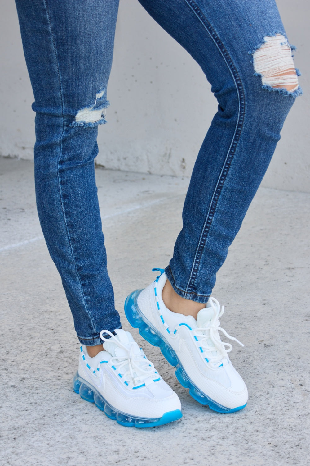 A person wearing ripped jeans and stylish Forever Link Lace-Up Air-Cushioned Athletic Shoes in white with blue accents stands confidently on a concrete surface.